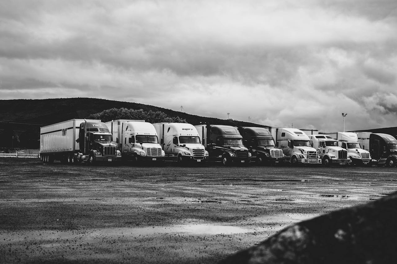 heros-img Row of parked semi trucks in a rainy lot, captured in a dramatic black and white setting.